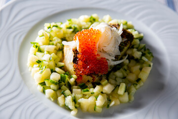 Chef pouring creamy sauce onto an elegant gourmet seafood appetizer with red caviar and chopped vegetables on a white plate.