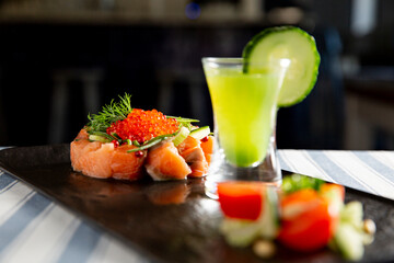 Gourmet salmon tartare with vibrant red caviar on a black plate. Served with fresh cucumber, dill, and a green shooter cocktail.