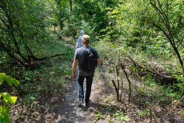 Two people and a dog hike through a lush, sun-dappled forest path, enjoying nature and outdoor activity. A man with a backpack leads the way, surrounded by vibrant green foliage.