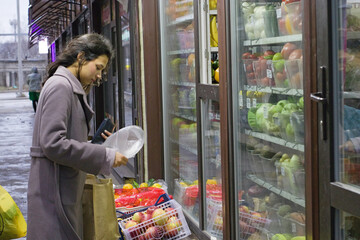 Photo of a young Indian woman in a winter coat selecting apples at an outdoor fruit stand in an urban setting. Useful for articles, blogs, markets, or healthy lifestyle promotions