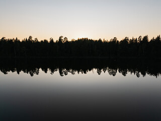 Aerial drone view of dark forest silhouette reflected in calm lake water during tranquil sunset in the Estonian countryside nature