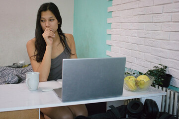 Young woman in a casual tank top sits at a desk with a laptop, mug, notebook, and a bowl of apples, looking thoughtful. Appropriate for remote work, studying, freelance, or home office themes.