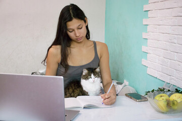 Young woman in a tank top sits at a white desk with a laptop, notebook, apples, and a cat on her lap, holding a pen and looking away. Suitable for study, pet, work from home, or cozy living stories.
