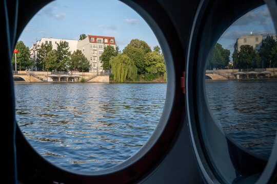 Urban view of Berlin waterfront seen through a circular window, showing reflection across the Spree River with calm housing architecture and a strong sense of tranquility