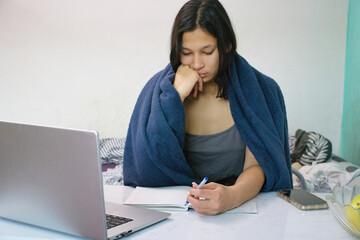 Young woman in a blue blanket studies at a desk using a laptop, with a dog on her lap, notebook, phone and fruit on the table. Good for distance learning, pets, or daily routines at home