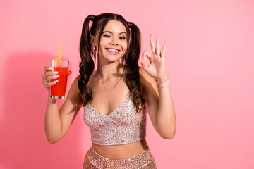 Smiling young woman with pink background holding a red drink and showing an ok gesture in a sparkly...