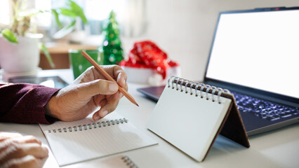close up shot, hand holding pencil taking note on blank calendar working with laptop, red hat, christmas tree for decoration on desk, christmas holidays concept