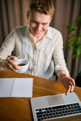 Smiling man working on laptop with coffee cup in hand at wooden desk. Concept of productivity support, workflow organization, daily planning, office lifestyle, and beverage-focused visual use.