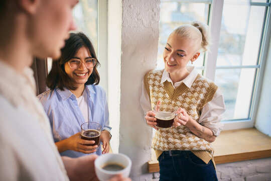 Coworkers laughing together while holding coffee near a bright window. Concept of positive team bonding, workday pauses, shared energy, and supportive office relationships.