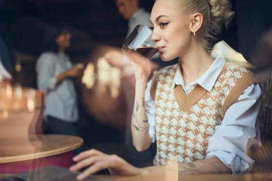 Woman sipping coffee while working in office with colleagues talking nearby. Concept of mindful coffee moments, hybrid work habits, light productivity, and lifestyle-focused workspace visuals.