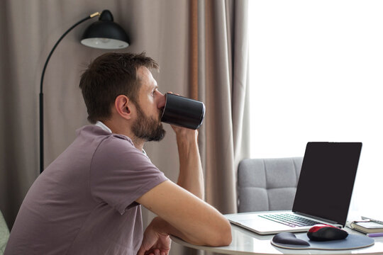 A man drinking coffee while sitting at a table with a laptop at home. Morning routine, remote work atmosphere, relaxed lifestyle and home office concept with natural daylight.