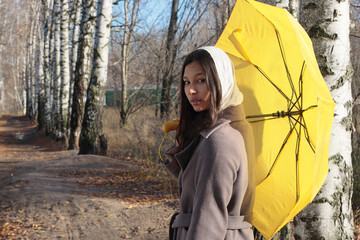 Young woman in a headscarf and brown coat stands on a forest path lined with birch trees, holding a yellow umbrella and looking at the camera. Suitable for autumn walk, beauty, seasonal content.