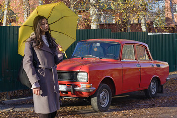 A young Indian woman strolls past a red vintage car, holding a yellow umbrella, side view