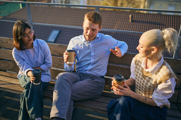 Coworkers sitting together on bench outdoors enjoying coffee and conversation. Concept of informal team dialogue, workplace culture, outdoor meetings, and relaxed professional interactions.