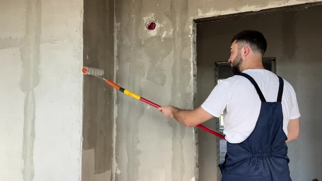 A bearded man in a work uniform is priming the walls after applying gypsum plaster. 