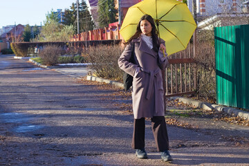 Young woman in a brown coat and headscarf walks with a yellow umbrella along a paved street, passing by houses and trees on a sunny day. Great for city walk, outdoor activity, or lifestyle stories
