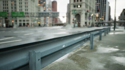 A quiet city intersection features a gray guardrail along the sidewalk, glistening from recent rain.