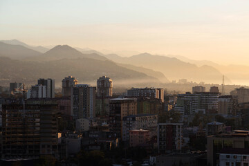 Obraz premium Sunset cityscape of Batumi with residential buildings, soft haze, layered mountain silhouettes and warm evening light. Scenic urban landscape of a coastal city surrounded by hills and nature.