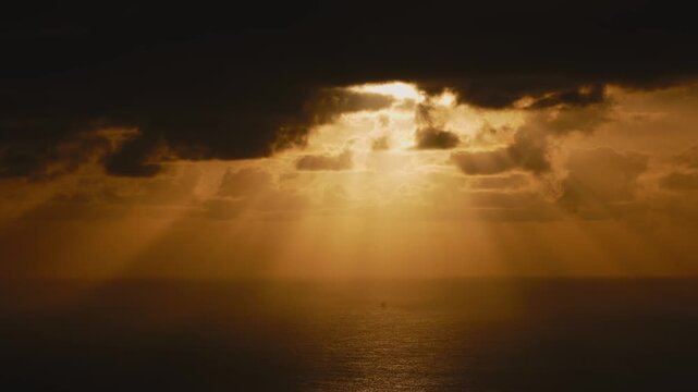 Golden rays of sunlight break through dark clouds over the ocean, illuminating the water surface, creating a serene atmosphere.
