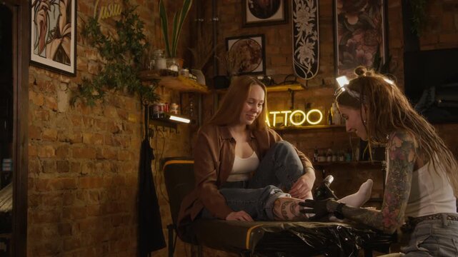 Handheld shot of professional young Caucasian female tattooist with dreadlock hairstyle using tattoo machine while working with female customer in cozy modern studio