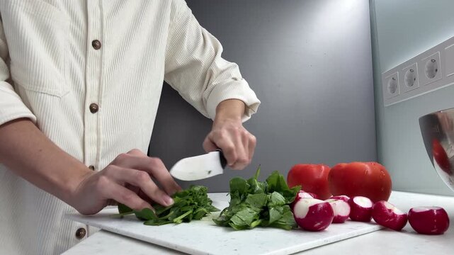 Woman Preparing Fresh Salad at Home &ndash; Cutting Spinach Leaves on Kitchen Counter with Knife, Radishes and Tomatoes Nearby, Healthy Cooking Scene
