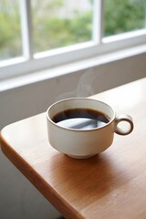 A steaming cup of coffee sits peacefully by a window on a wooden table top surface
