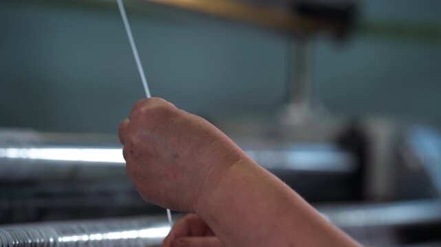 A hand carefully separates white textile threads across smooth polished metal rollers of a weaving machine, highlighting precise manual control, soft lighting and calm workshop motion