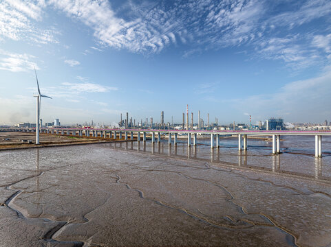 A large petrochemical plant and wind turbine on the coast with a long bridge over the mudflats. - Powered by Adobe