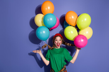 Joyful young girl with colorful balloons in bright purple studio celebrates fun fashion moment for...