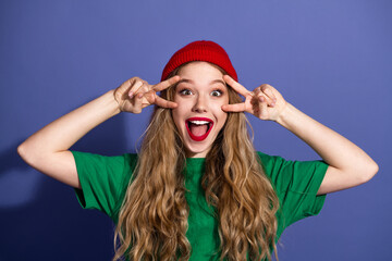 Young woman with red beanie and green shirt making a playful peace sign and smiling wide against...