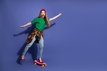 Fototapeta premium Young girl on a skateboard in a green shirt red beanie plaid shirt jeans and red heels smiling against a blue background casual stylish lifestyle fashion shot