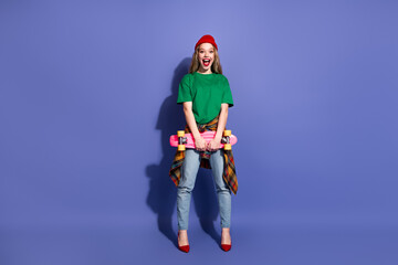 Young woman in green shirt red beanie holding pink skateboard against blue background exuding...