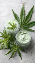 Overhead close-up shows two glass jars filled with soothing hemp-derived skincare cream, decorated with fresh green cannabis leaves, on a textured gray surface.