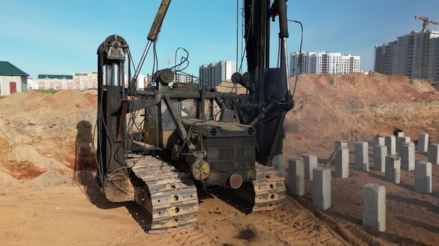 Powerful diesel piling rig operates to install concrete piles on a construction site surrounded by buildings under a bright sky.