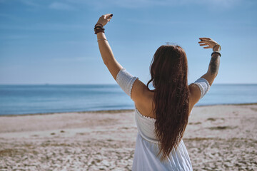 Rearview of young woman in white summer dress stands alone on sandy beach with her arms lifted toward blue sky, enjoy summer vacation and view, welcoming ocean. Freedom, balance and peace concept