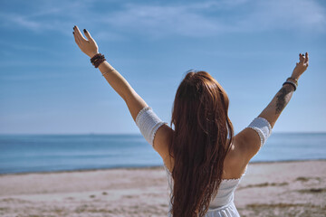 Rear view of young woman in white dress stands on sunny beach with her arms raised toward sky, feeling freedom and tranquillity of ocean. Deep connection with nature and inner balance concept