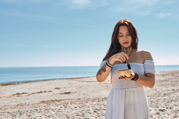 Young 20s Caucasian woman in white dress stands on sandy sunny beach holding Tibetan singing bowl, focusing on sound meditation outdoors. Concept of inner peace and harmony with nature