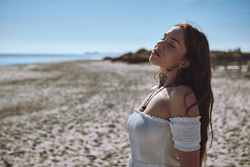 Young woman in white dress stands on beach with closed eyes, breathes deeply and enjoying moment of mindfulness outdoors, sunlight and sea breeze evoke calmness, balance, and emotional harmony concept