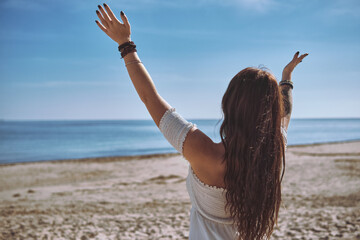 Rear view of young woman raises her hands in serene gesture of mindfulness while facing calm blue ocean on the beach. Freedom, spiritual renewal by ocean