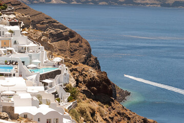 Santorini coast with white houses cascading on rocky cliffs toward the deep blue sea crossed by a speedboat.