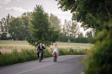 Obraz premium Couple riding bicycles along a scenic country road, surrounded by lush greenery and fields, enjoying a sunny day outdoors, capturing the essence of leisure and connection