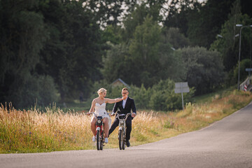 Obraz premium Couple riding bicycles on a scenic road, with lush greenery and golden grass surrounding them, enjoying a joyful moment in a serene outdoor setting