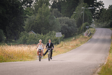 Couple riding bicycles along a scenic rural road, surrounded by lush greenery and golden fields, enjoying a sunny day, capturing the essence of love and adventure