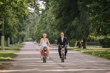 Fototapeta premium Couple riding bicycles on a tree-lined path, showcasing joy and connection, dressed in formal and casual attire, capturing a moment of togetherness and celebration