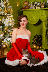 Portrait of a young attractive woman in a red Christmas dress and long gloves posing sitting with a cat in her hands against the background of a Christmas tree and decorations.