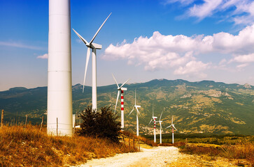 Wind turbines lined along a dirt road in a sunny mountain landscape, blending nature with clean energy.