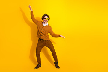 Young stylish man dancing in a yellow backdrop wearing a brown cardigan and sunglasses
