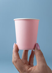 Female hand with disposable pink paper cup against light blue closeup, mockup, copy space