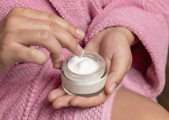 Young woman in pink bathrobe touching cream with finger and holding a cosmetic jar closeup