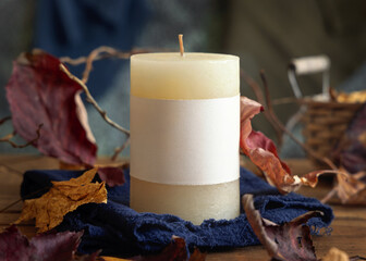 Candle with a label near autumn dried leaves closeup,  fall mockup in blue, red and orange